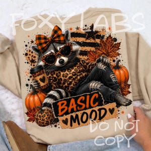 May include: A beige sweatshirt featuring a graphic of a raccoon wearing a leopard print sweater, orange plaid bow, and sunglasses. The raccoon holds a coffee cup near pumpkins and autumn leaves. The text reads BASIC MOOD with heart accents.