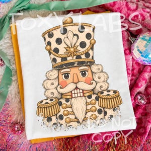 May include: A white t-shirt featuring a graphic illustration of a nutcracker soldier with a cream and black polka dot uniform, gold braided trim, and a tall hat. The design includes a white beard, moustache, and a star emblem.