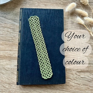 May include: A green, intricately designed bookmark rests on a dark blue book. The bookmark features a detailed, openwork pattern. The text "Your choice of colour" is displayed on the right side of the book. The background is a light brown wooden surface.