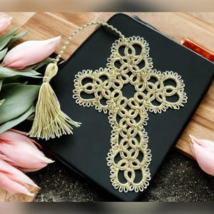May include: A gold crochet cross bookmark with a tassel. The bookmark is laying on a black book cover. The book cover is on a wooden surface.