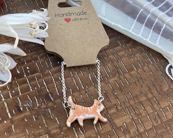 May include: A handmade necklace with a small orange and white cat pendant. The cat charm is attached to a silver chain. The necklace is displayed on a wooden surface with a tag that reads "Handmade with love."