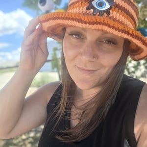 May include: A crochet bucket hat featuring horizontal stripes in shades of orange, brown, and peach. The hat is adorned with a prominent black, white, and blue evil eye patch centered on the front, showcasing a detailed iris and pupil design.