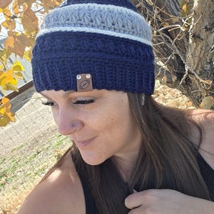 May include: A navy blue, gray, and white striped crochet beanie with a cream-colored pom-pom. The beanie has a brown leather tag with a football graphic. The hat is handmade.