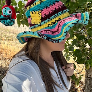 Op de afbeelding: Een kleurrijke, handgehaakte hoed met een brede rand. De hoed heeft een patchwork-ontwerp met verschillende kleuren, waaronder geel, roze, teal en bordeaux, en is versierd met zwarte kruissteekdetails. Een bijpassend gehaakt ornament met een karakter is ook zichtbaar.