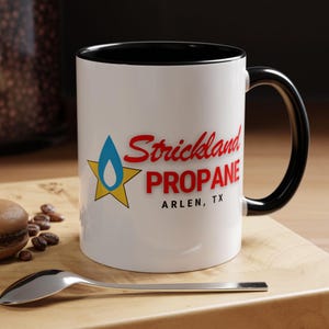 May include: White ceramic mug with a black rim and handle. The mug features a red and blue logo that says "Strickland Propane" with the text "Arlen, TX" underneath.