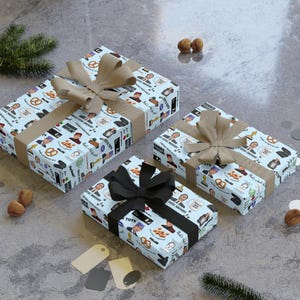 Könnte beinhalten: Drei rechteckige Geschenkboxen, eingewickelt in hellblaues Papier mit einem wiederkehrenden Muster aus verschiedenen Symbolen und Texten, darunter Dunder Mifflin, Brezeln und Charakterillustrationen. Zwei Boxen haben beigefarbene Schleifen, eine hat eine schwarze Schleife.