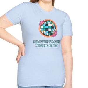 May include: Light blue t-shirt featuring a disco ball graphic with a rainbow, flowers, and the text "HOOTIE TOOTIE DISCO CUTIE". The graphic is in shades of teal, black, white, pink, and yellow. The shirt has short sleeves and a crew neck.