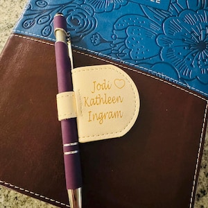 Puede incluir: Primer plano de una Biblia con una cubierta azul con diseño floral y el texto "The Woman's Study Bible". Un bolígrafo morado descansa sobre un portalápices crema con el nombre "Jodi Kathleen Ingram" en letras doradas.