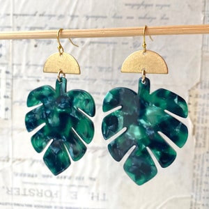 May include: A pair of dangle earrings featuring a tropical leaf design. The earrings have a gold-colored half-circle top and a green and blue marbled leaf pendant. The earrings are hanging on a wooden dowel against a vintage paper background.