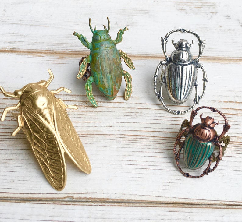 May include: Four insect-themed brooches in gold, silver, and green tones. The brooches include a cicada, a scarab beetle, and two other beetle designs. The brooches are displayed on a white wooden surface.