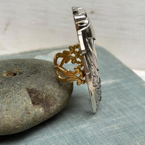 May include: A silver-toned ring with an ornate, rectangular design. The ring features a gold-colored filigree band. The ring is displayed on a gray stone, showcasing its intricate details and contrasting colors.