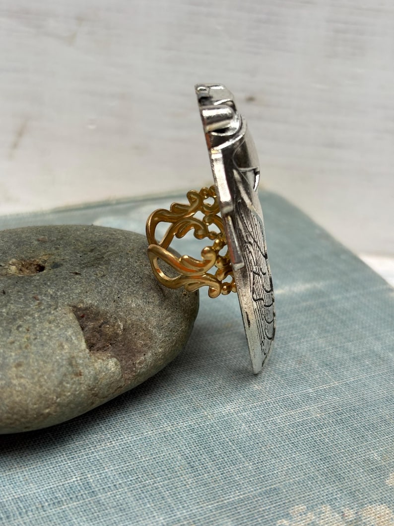 May include: A silver-toned ring with an ornate, rectangular design. The ring features a gold-colored filigree band. The ring is displayed on a gray stone, showcasing its intricate details and contrasting colors.