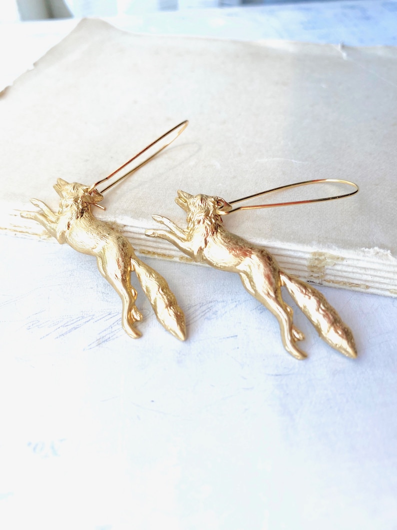 Puede incluir: Un par de pendientes de zorro dorados. Los zorros est&aacute;n representados en una pose de salto con las colas enroscadas detr&aacute;s de ellos. Los pendientes est&aacute;n hechos de metal y tienen un dise&ntilde;o simple y elegante.
