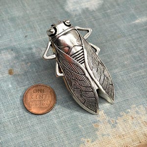 May include: A silver-colored cicada brooch with detailed wing and body textures. A copper one-cent coin is next to the brooch for size comparison. The brooch is on a blue-gray textured surface.