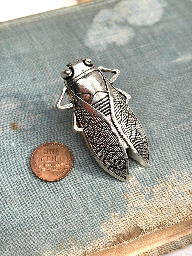May include: A silver-colored cicada brooch with detailed wing and body textures. A copper one-cent coin is next to the brooch for size comparison. The brooch is on a blue-gray textured surface.