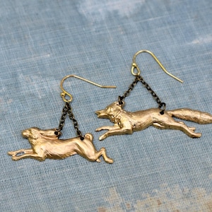 May include: A pair of gold-colored earrings featuring a fox and a hare in mid-stride. The fox and hare are suspended from a gold-colored hook by a short, dark chain. The earrings are set against a blue background.