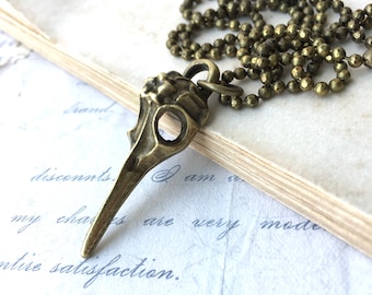 May include: A bronze-colored pendant necklace featuring a bird skull design. The pendant is intricately detailed with an open beak and eye socket. The necklace has a ball chain and is set against a backdrop of vintage paper with handwritten script.
