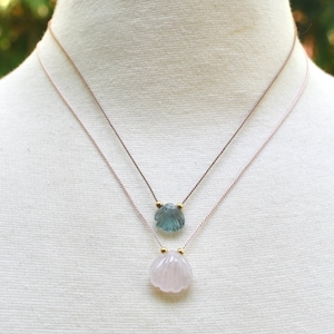 May include: Two delicate necklaces with seashell pendants. The top pendant is a light blue color, and the bottom pendant is a soft pink. Both pendants are strung on thin, light pink cords with gold accents. The necklaces are displayed on a white mannequin.