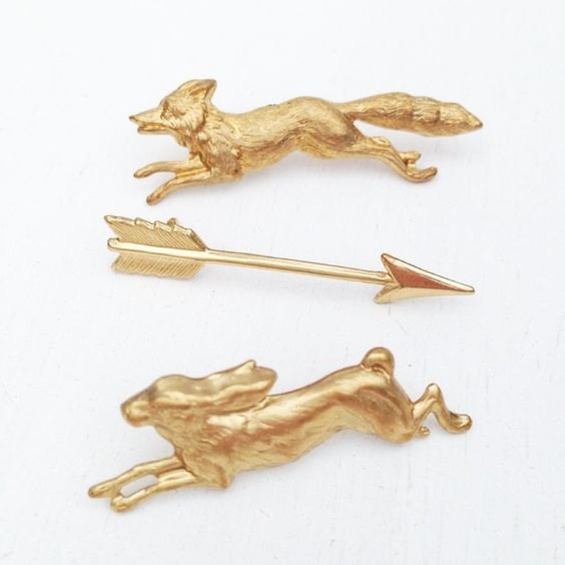 Puede incluir: Tres figuritas de animales de metal de color dorado: un zorro corriendo, una flecha apuntando hacia la derecha y un conejo corriendo. Las figuritas est&aacute;n sobre un fondo blanco.