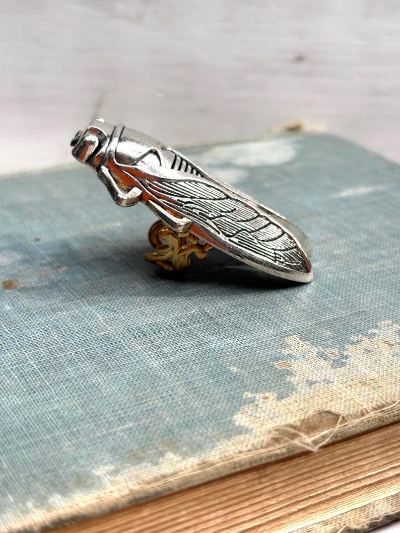 May include: A silver-colored cicada brooch with detailed wing and body features. The insect-shaped pin has a gold-colored base and is set against a vintage book. This unique jewelry piece is a nature-inspired accessory.