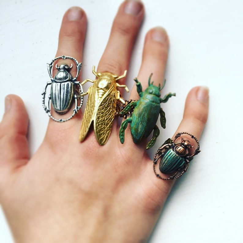 May include: Four insect-themed rings in silver, gold, and green tones. The rings feature detailed designs of beetles and a cicada, showcasing intricate textures and shapes. The rings are worn on a hand against a white background.
