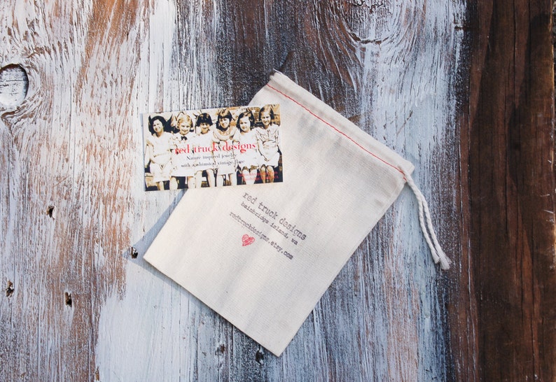May include: A small, off-white drawstring bag and a business card rest on a weathered wooden surface. The card features a vintage photo and the text "red truck designs." The bag has red stitching and the brand name printed on it.