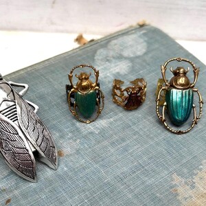May include: A collection of insect-themed jewelry, including a silver cicada brooch and several rings. The rings feature beetle designs with green and gold accents. The jewelry is displayed on a vintage blue book.