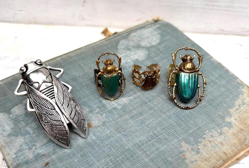 May include: A collection of insect-themed jewelry, including a silver cicada brooch and several rings. The rings feature beetle designs with green and gold accents. The jewelry is displayed on a vintage blue book.