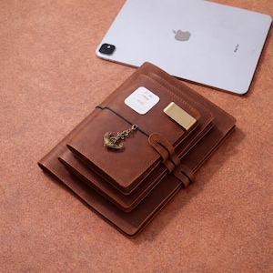 May include: A stack of three brown leather journals with a brass clip and an anchor charm. A white tablet with the Apple logo is in the background. The journals have a strap closure and a small sticker.
