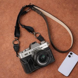 May include: A silver and black Fujifilm X-T30 camera with a black lens, encased in a black leather-textured half case. The camera features a black and brown leather neck strap with metal rings and a light purple smartphone positioned nearby.