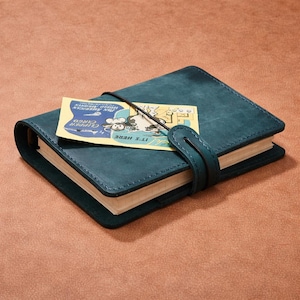 Peut inclure: Journal en cuir bleu canard avec une fermeture à lanière et une petite étiquette. L'étiquette présente une illustration vintage et le texte "PAN AMERICAN CLIPPER CARGO". Le journal est fermé par un élastique noir.