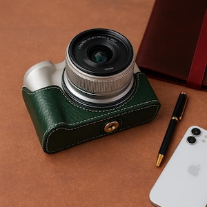 Op de afbeelding: Een zilveren en groene camera met een lens, in een groene leren hoes. Een zwarte pen en een witte smartphone zijn ook zichtbaar. De camera staat op een bruin oppervlak.