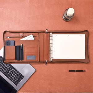 May include: A brown leather portfolio with a zipper closure, open to reveal a notepad and binder rings. The portfolio has pockets for a phone, cards, and a pen. A laptop and pen are also on the table.