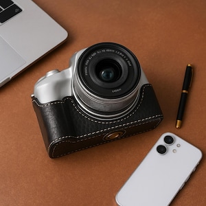 May include: A silver and black digital camera with a black leather case, a black lens, and a white smartphone are on a brown surface. A silver laptop and a gold and black pen are also present.