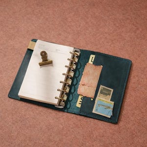 Puede incluir: Diario de cuero verde azulado abierto con papel rayado, un clip dorado y una encuadernación en espiral de metal. El diario está abierto para revelar un bolsillo con sellos antiguos y un trozo de papel rasgado.