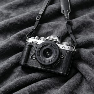 May include: A silver and black Nikon Zfc mirrorless camera with a black lens and a textured black leather-like half case. The camera features a silver top plate with dials and a black neck strap with white stitching attached to the sides.