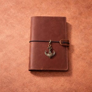 May include: A brown leather journal with an anchor charm. The journal is closed with a black elastic band and a leather strap. The anchor charm is a bronze color and hangs from the elastic band.