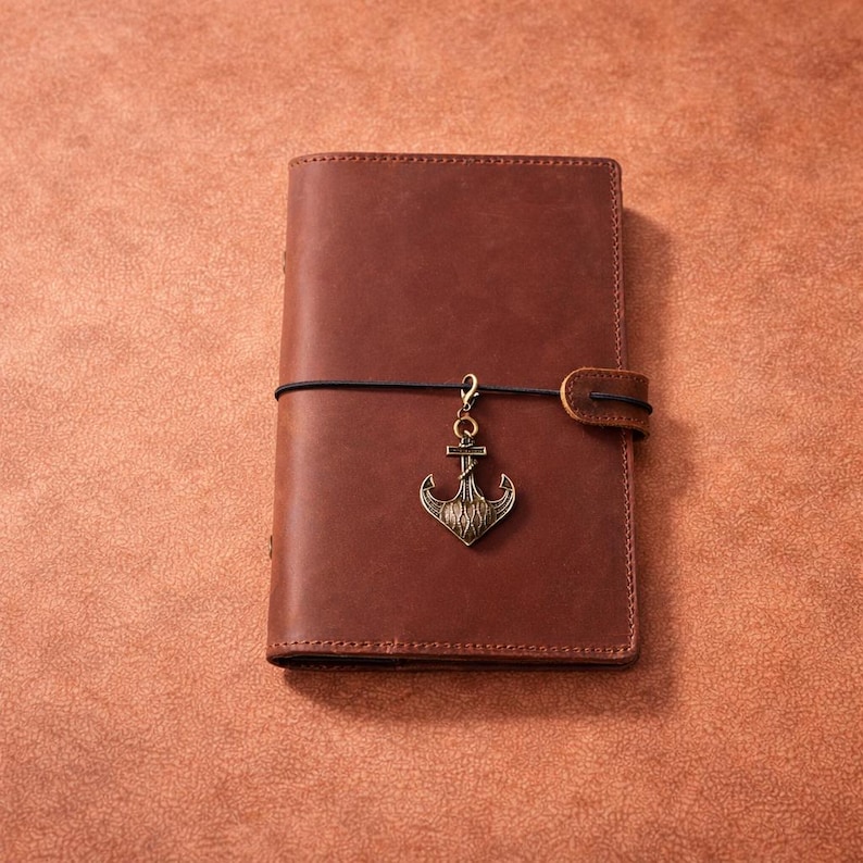 May include: A brown leather journal with an anchor charm. The journal is closed with a black elastic band and a leather strap. The anchor charm is a bronze color and hangs from the elastic band.