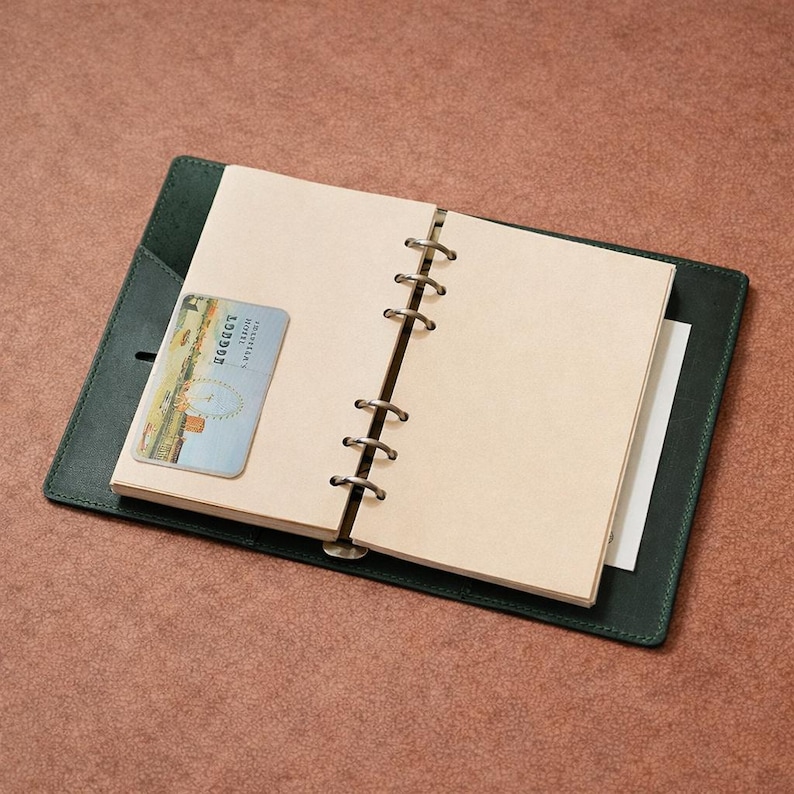 May include: An open, dark green leather journal with a light brown interior and a small card featuring a London image. The journal is on a brown surface. The journal has a ring binder mechanism.