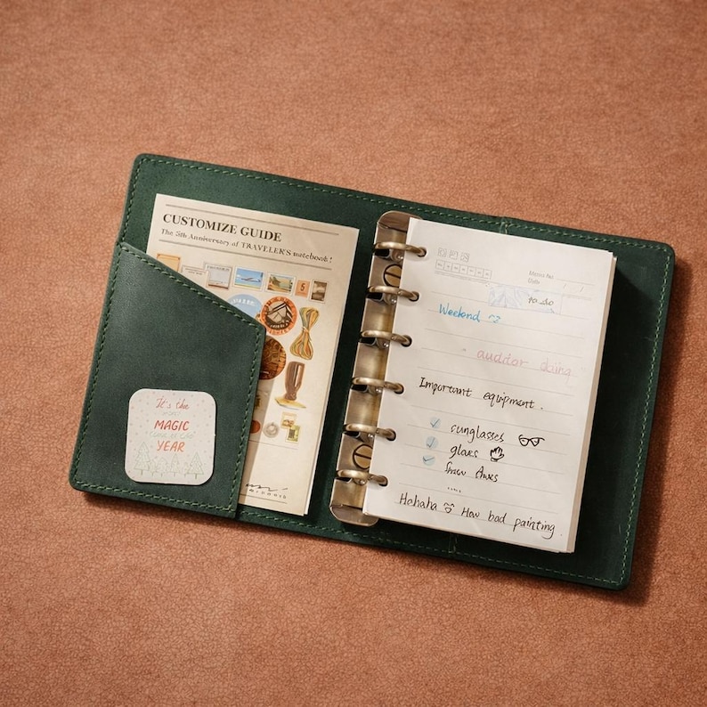 May include: Open green leather notebook with a metal binder. The notebook contains a "Customize Guide" and a lined paper with handwritten notes. A small square sticker reads "It's the MAGIC YEAR".
