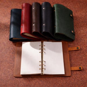 Puede incluir: Una colección de diarios de cuero en varios colores, incluyendo azul marino, rojo, marrón, negro y verde. Un diario está abierto, revelando páginas blancas en blanco y una encuadernación de anillas. Los diarios están dispuestos sobre una superficie marrón.