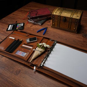 May include: A tan leather portfolio organizer featuring a six-ring metal binder mechanism, multiple interior card slots, a zippered pocket, and a pen holder. The open organizer contains blank white paper sheets and a small decorative paper cone with flowers.