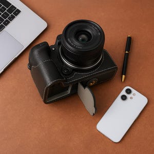 May include: Black leather camera case with white stitching, holding a camera with a lens. A black pen and a white smartphone are next to the camera. The items are on a brown surface.