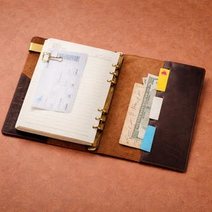 May include: Open brown leather journal with lined pages, a gold-colored clip, and a boarding pass. The journal has a pocket with cash and sticky notes. The journal is open on a brown surface.