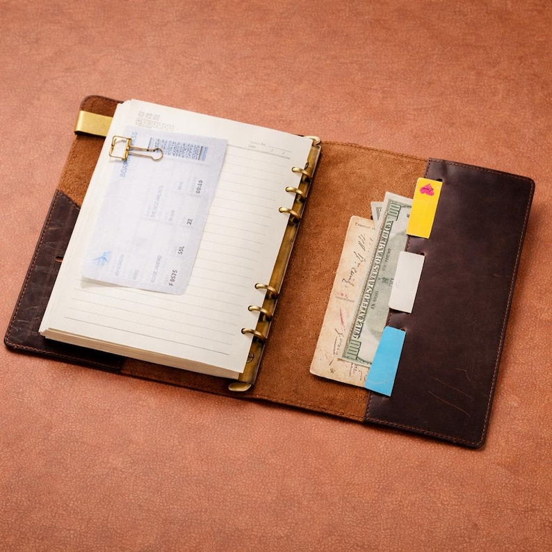 May include: Open brown leather journal with lined pages, a gold-colored clip, and a boarding pass. The journal has a pocket with cash and sticky notes. The journal is open on a brown surface.