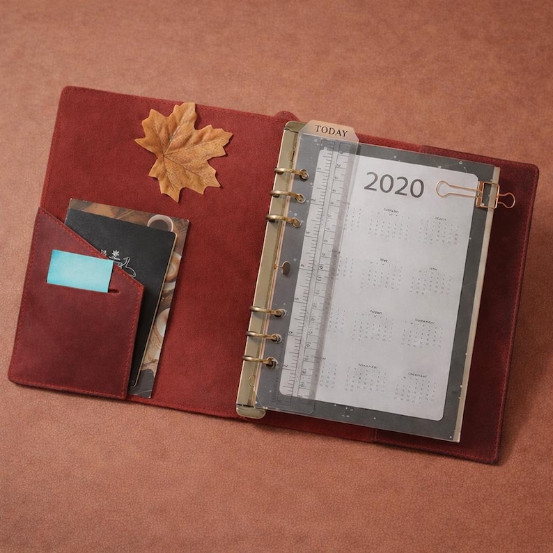 May include: Open burgundy leather planner with gold-colored rings, a 2020 calendar, and a clear ruler. A small pocket holds cards and a blue sticky note. A decorative leaf rests on the cover.