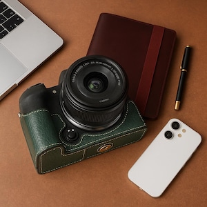 May include: A digital camera with a black lens and a green leather case sits on a brown surface. A white smartphone, a burgundy notebook, a black pen, and a laptop are also visible.