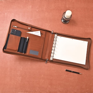 May include: Open brown leather portfolio with multiple compartments, including a phone pocket, card slots, and a zippered section. It features a binder mechanism and a pen holder. A black pen and a smartphone are visible.