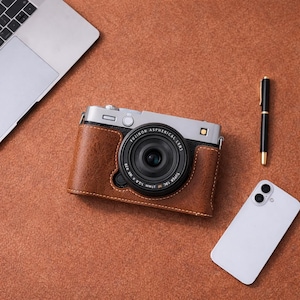 Könnte beinhalten: Eine silberne und schwarze Digitalkamera mit einer braunen Lederhülle, ein schwarzer Stift, ein weißes Smartphone und ein Laptop sind auf einer braunen Oberfläche angeordnet. Das Kameraobjektiv ist zentriert, und die Hülle hat sichtbare Nähte.