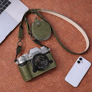 May include: A green leather camera case with a matching strap and small round pouch. The camera is silver and black, with a lens and the Fujifilm logo. A white smartphone and laptop are also in the image.